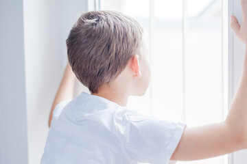 Little boy 7 years old in a white T-shirt in a bright room looks out the window. Emotions.