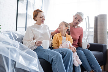 cheerful girl sitting on couch near same sex parents in new apartment