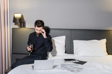 bearded man holding glass of whiskey and looking at laptop in hotel