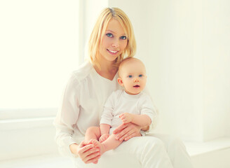 Happy smiling young mom playing with her little baby together in white room at home