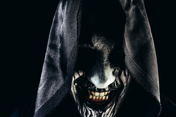 Horror photo of a male monster reaper smiling face in hood on dark background.