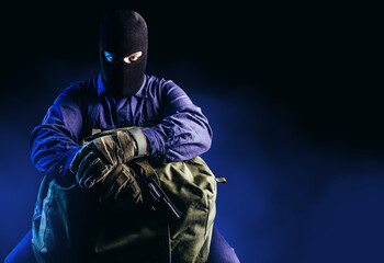 Photo of robber in mask, overalls, gloves, gun sitting and holding bag on dark blue background. - obrazy, fototapety, plakaty