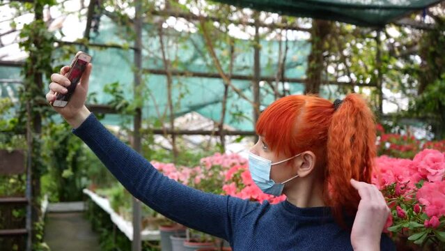 a young red-haired girl in a greenhouse in a medical mask takes pictures of herself on the phone against the background of azaleas flowers. slow motion video close-up