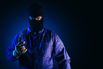 Photo of robber in mask, overalls, gloves and gun standing on dark blue background.