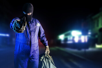 Photo of robber in mask, overalls, gloves, bag and gun standing front view and aiming on night...
