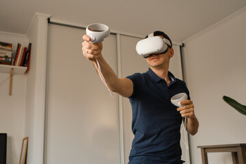 Young man in casual clothes plays virtual games at home. Man in virtual reality VR helmet compensates lack of movement.