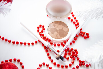 A white pillbox with powder and bronzer lies on a white background, thin makeup brushes and bright red beads lie in front. High quality photo
