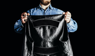 Photo of biker man in jeans shirt holding leather vest with patch colors on black background.