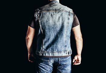 Back view photo of man in jeans shirt and denim biker vest standing on black background.
