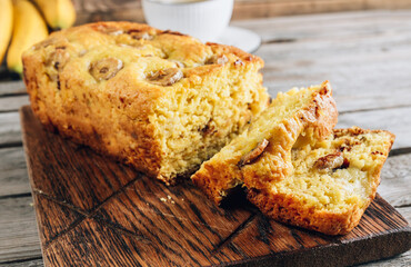 Loaf of banana bread on a wooden cutting board with cup of tea on rustic wooden table.
