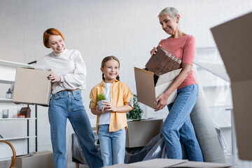 lesbian women holding cardboard boxes near happy child with flowerpot in new apartment