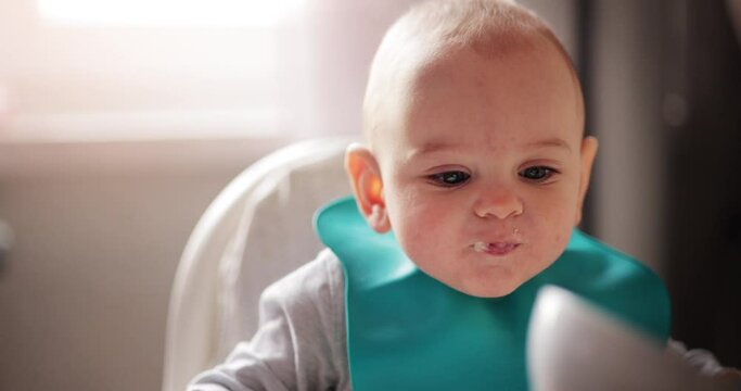 Happy toddler baby visibly enjoying the taste of healthy solid food, being fed with a spoon while sitting in a high chair, slow motion 60 FPS