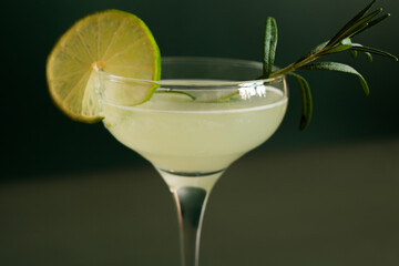 Citrus alcoholic cocktail. Close up of drinking glass of daiquiri cocktail decorated lime and rosemary on dark green background. Selective focus.