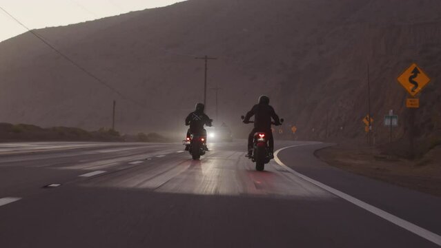 Men ride their fast bikes during twilight over a coastal town with rocky cliff behind. Scenic view of the bikers driving motorcycles towards oncoming cars. High quality 4k footage
