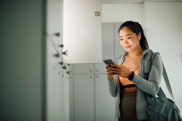 Smiling Asian athletic woman texting on smart phone at gym's locker room.