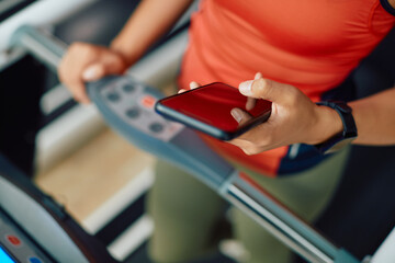 Close up of sportswoman texting on cell phone while practicing on treadmill in gym.