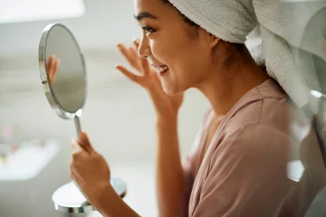 Poster Smiling Asian woman takes care of her facial skin and applying moisturizer in bathroom. © Drazen