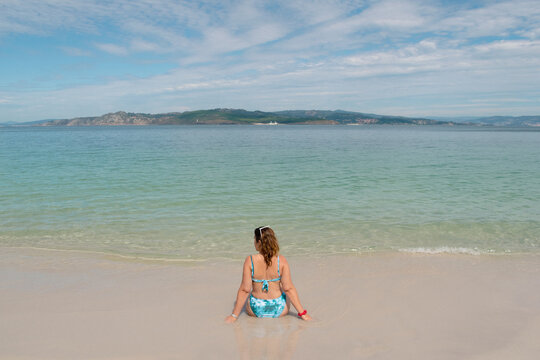 Rear view of a woman in a bikini sitting on beach by water's edge, Spain