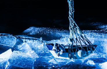 Photo of blue toned miniature sailboat standing on cracked ice surface on dark background.
