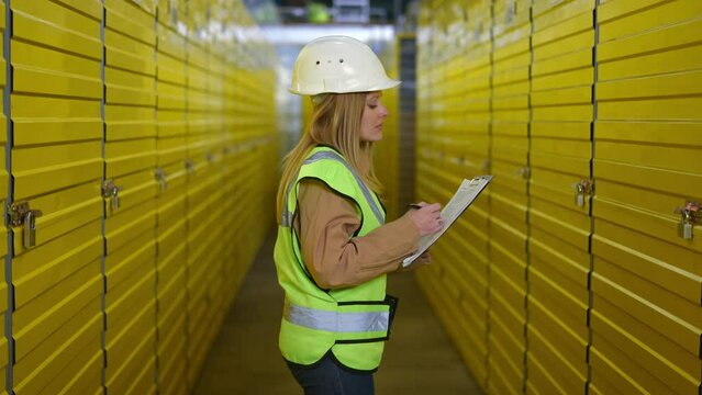 Back view young female employee in uniform walking along rows of yellow lockers in warehouse filling in paperwork. Tracking shot of Caucasian woman strolling in industrial storage inspecting goods
