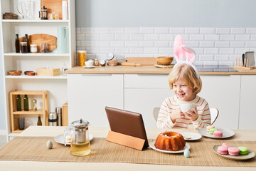 Fototapeta na wymiar Portrait of cute boy wearing bunny ears while enjoying breakfast on Easter morning and using tablet in kitchen, copy space