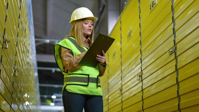 Portrait of thoughtful young woman in uniform standing in warehouse filling in paperwork. Concentrated Caucasian female freight agent indoors in storage writing in documents thinking