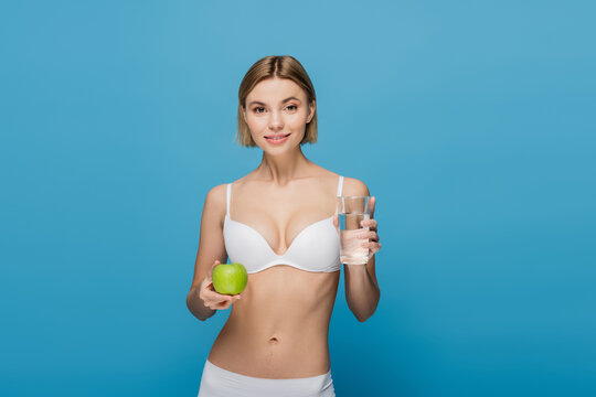 smiling young woman in white bra holding green apple and glass of water isolated on blue
