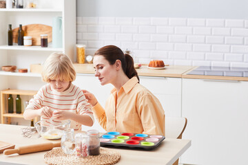 Fototapeta premium Portrait of young woman baking cupcakes with cute little boy helping mom in kitchen, copy space