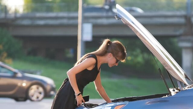 Helpless woman standing near her car with open hood takling on mobile phone calling road service for help. Young female driver having trouble with vehicle