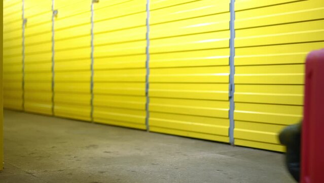 Legs of unrecognizable young Caucasian woman walking with rolling suitcase along row of yellow storage rooms doors. Female tenant with luggage passing in self storage indoors