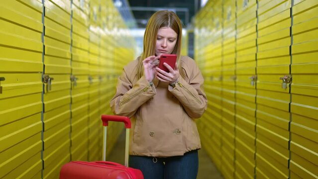 Medium shot doubtful young woman using mobile app on smartphone standing with luggage in self service storage. Portrait of Caucasian unsure lady searching lockers using phone application