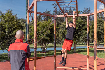 A young Latino man crosses the monkey bar in his exercise routine while his instructor watches him