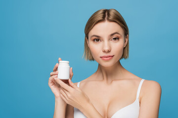 blonde young woman in white bra holding bottle with vitamins isolated on blue