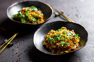 chicken wok noodles with vegetables and sesame and wok with fried beef and vegetables on black bowls on dark stone table side view with selective focus