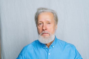 Close-up portrait of serious gray-haired senior man unemotionally looking at camera on white...