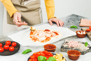 Hands of a caucasian teenage girl in an apron smear tomato sauce on a heart-shaped pizza dough.