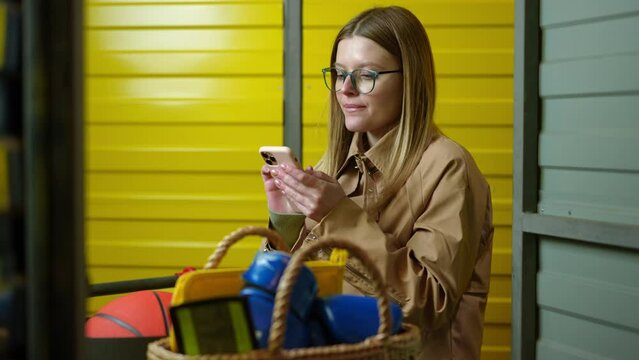 Young woman messaging online in social media in smartphone app standing with belongings in self storage. Caucasian beautiful female tenant in yellow warehouse indoors