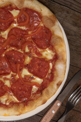 Pizza lista para comer con salsa roja, queso mozzarella, peperoni y pimiento en un plato blanco sobre un fondo de madera junto a un cuchillo y un tenedor