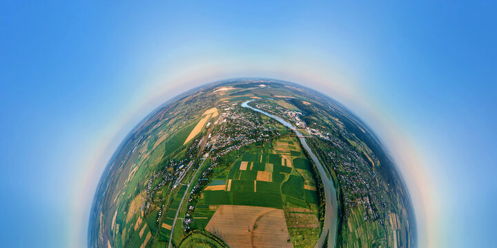 Aerial view from high altitude of little planet earth with small village houses and distant green cultivated agricultural fields with growing crops on bright summer evening - Powered by Adobe