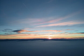 Aerial view of colorful sunset over white dense foggy clouds cover with distant dark silhouettes of mountain hills on horizon