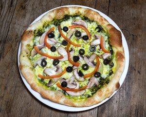 Pizza con salsa pesto, queso mozzarella, champiñon, aceitunas y pimientos en un plato blanco sobre un fondo de madera
