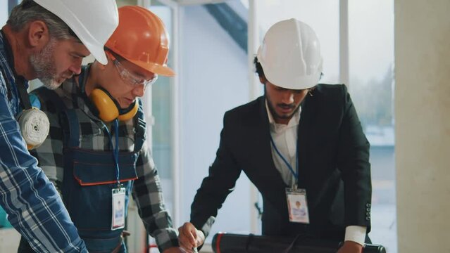 Close up architect man in suit works with engineers or builders in hardhats checking plan details in building. Talking, discussing, drawing on blueprint. Slow motion