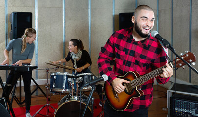 Rehearsal of music band. Cheerful positive smiling guy guitar player and singer practicing with band members in recording studio.