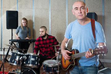 Glad male singer with guitar rehearsing with band of drummer and keyboardist before public performance