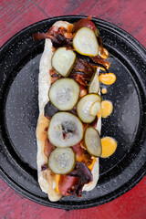 American food. Fusion. Closeup view of a gourmet hot dog with crispy bacon, cheddar cheese and cucumber slices, presente don a black dish on the red wooden table.