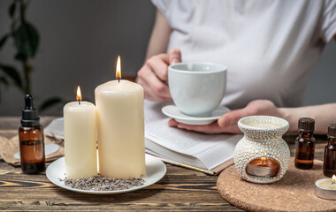 Fototapeta na wymiar Woman is reading a book and drinking tea in atmosphere of harmony and relaxation. Aroma lamp with essential oils and burning candles on the wooden table