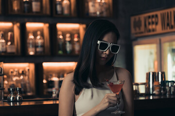 Asian woman drinking a cocktail at a bar while wearing sunglasses.