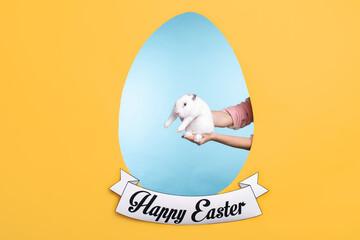 Cute white fluffy bunny on hands of young woman on Easter greeting card with egg shape in the center and yellow copyspace around