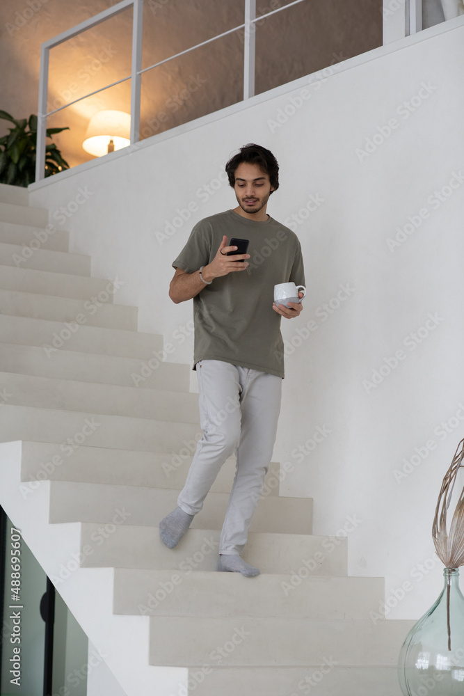 Poster contemporary guy with mug of tea and smartphone texting in mobile gadget while moving down along whi