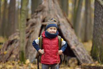 Little boy scout during hiking in autumn forest. Behind the child is teepee hut. Adventure, scouting and hiking tourism for kids.